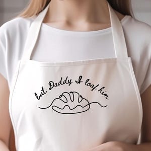 May include: Off-white apron with a black line drawing of a loaf of bread and the text "but Daddy I loaf him". The apron has a pocket and neck and waist straps. The person is wearing a white t-shirt.