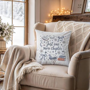 May include: A white square throw pillow with the text "Just one more chapter" in a whimsical font, surrounded by illustrations of books, glasses, and bows. The pillow is on a beige armchair.