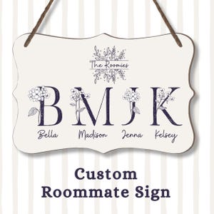 May include: A custom roommate sign with a cream background and a decorative border. The sign features the text "The Roomies" above the initials B, M, J, and K, each adorned with floral accents and the names Bella, Madison, Jenna, and Kelsey.