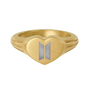 May include: A gold-colored ring with a heart-shaped face. The heart features a silver-colored logo. The ring has a brushed finish and a smooth band. The ring is a piece of jewelry.