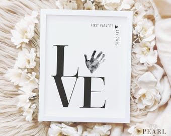 May include: A white framed print with the word "LOVE" in black text. The letter "O" is replaced with a black handprint. The text "First Father's Day 2025" is in the top right corner.
