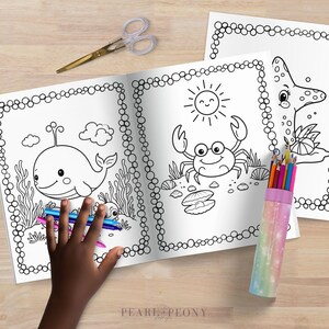 Puede incluir: Libro para colorear abierto con ilustraciones de vida marina, incluyendo una ballena, un cangrejo y una estrella de mar. Una mano sostiene l&aacute;pices de colores y unas tijeras est&aacute;n cerca. Las p&aacute;ginas tienen un borde punteado. Sobre una superficie de madera.
