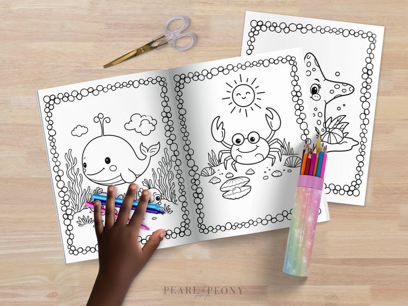 Puede incluir: Libro para colorear abierto con ilustraciones de vida marina, incluyendo una ballena, un cangrejo y una estrella de mar. Una mano sostiene l&aacute;pices de colores y unas tijeras est&aacute;n cerca. Las p&aacute;ginas tienen un borde punteado. Sobre una superficie de madera.