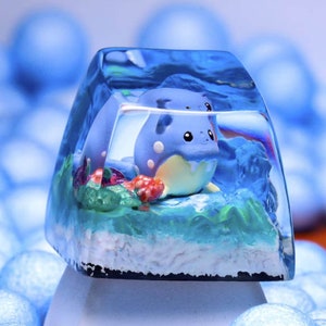 Puede incluir: Un tapón de tecla de resina azul y blanco con una pequeña figura azul y blanca de un personaje de Pokemon en su interior. El tapón de tecla está sobre una base blanca y rodeado de bolitas de embalaje azules.