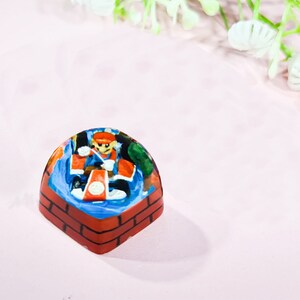 May include: A hand-painted resin keycap featuring a character in a red and blue outfit, holding a weapon, standing on a brick platform. The keycap is a dome shape with a clear resin top.