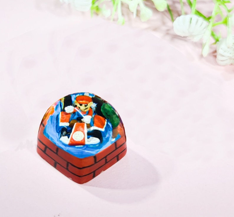 May include: A hand-painted resin keycap featuring a character in a red and blue outfit, holding a weapon, standing on a brick platform. The keycap is a dome shape with a clear resin top.