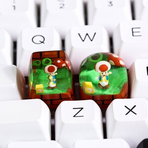 May include: Two clear plastic keycaps with a green, red, and brown mushroom-themed design. Each keycap features a small figurine of a mushroom character with a question mark block.