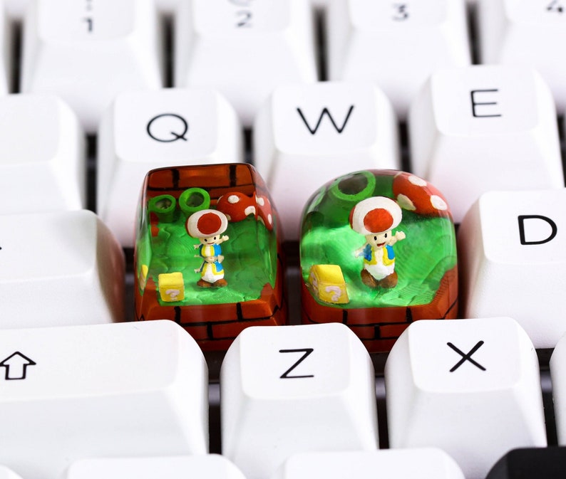 May include: Two clear plastic keycaps with a green, red, and brown mushroom-themed design. Each keycap features a small figurine of a mushroom character with a question mark block.