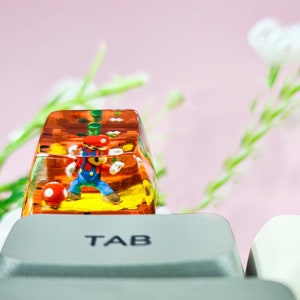 May include: A clear resin keycap with a small figurine of a video game character inside. The keycap is labeled "TAB" and is sitting on a white keyboard.