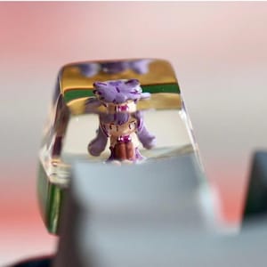 May include: A purple and white figurine of a character from a popular video game is encased in a clear resin keycap.