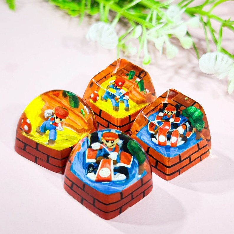May include: Four resin keycaps featuring different characters from the Super Mario Bros. video game series. Each keycap is a dome shape with a brick border and a clear resin top. The characters are Mario, Luigi, Toad, and a Goomba.
