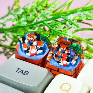 May include: Two resin keycaps featuring a cartoon character in a red racing suit driving a kart. The keycaps are red, orange, blue, and green with a brick-like design. The keycaps are on a keyboard with the keys "TAB" and "Q" visible.