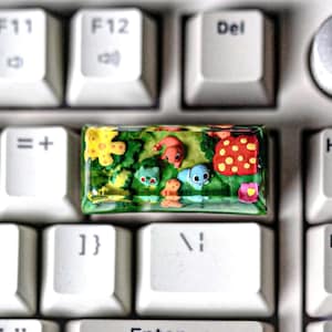 May include: A colorful keyboard keycap with a green, yellow, and red floral design. The keycap features a variety of small, cartoon-like characters, including a flower, a butterfly, and a ladybug.