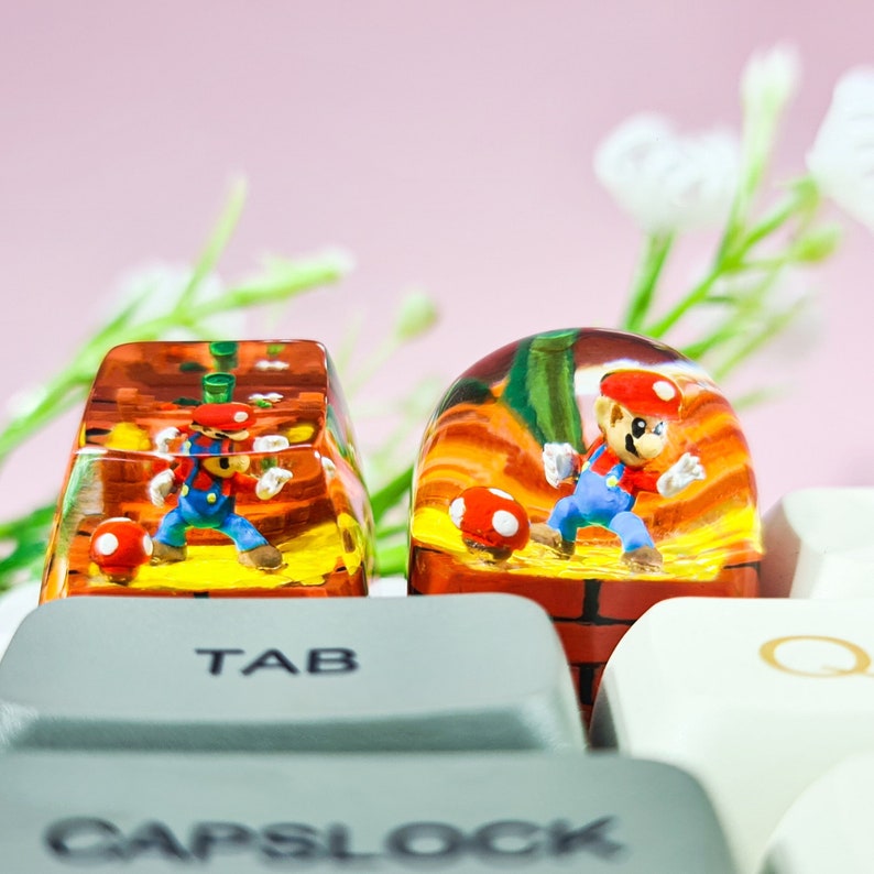 May include: Two clear resin keycaps featuring a miniature Mario character in a 3D scene. The keycaps are placed on a white keyboard with the words "TAB" and "CAPSLOCK" visible.