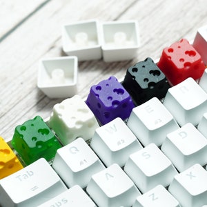 Könnte beinhalten: Eine weiße Tastatur mit bunten Tasten in Form von Käse. Die Tasten sind gelb, grün, weiß, lila, schwarz und rot.