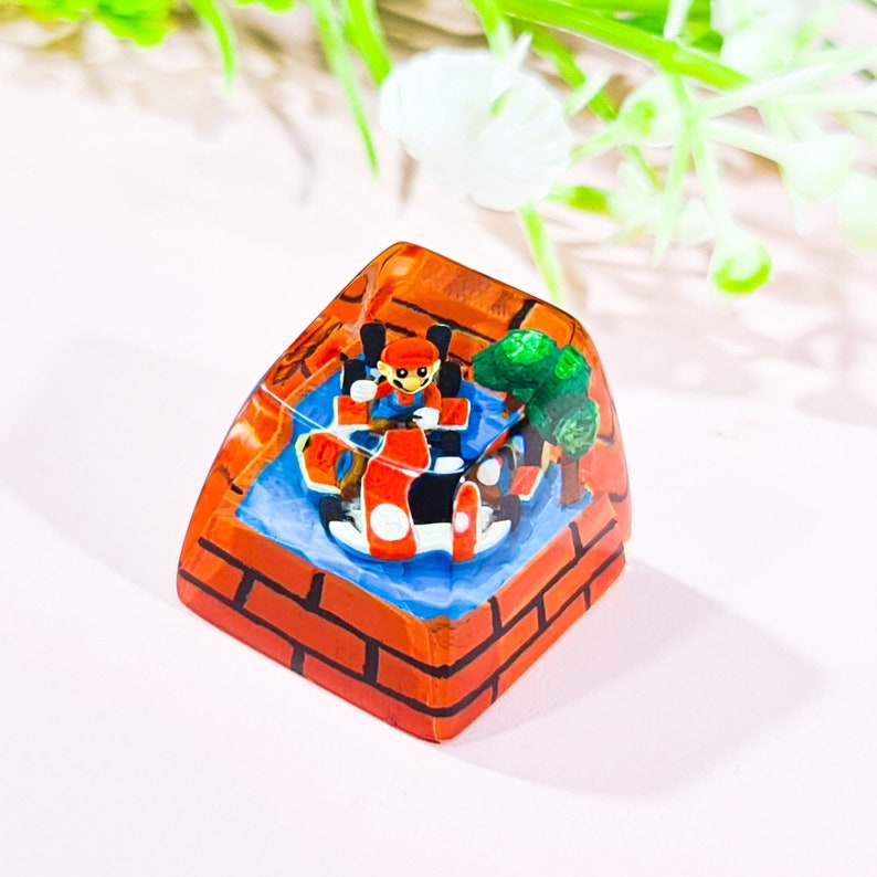 May include: A hand-painted resin keycap featuring a red and black go-kart with a figure of Mario driving. The keycap is shaped like a brick block and has a green bush on the side.