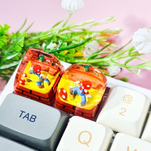 May include: Two resin keycaps featuring a video game character in a brick setting. The keycaps are on a white keyboard with the keys "TAB", "Q", "@", and "2" visible.