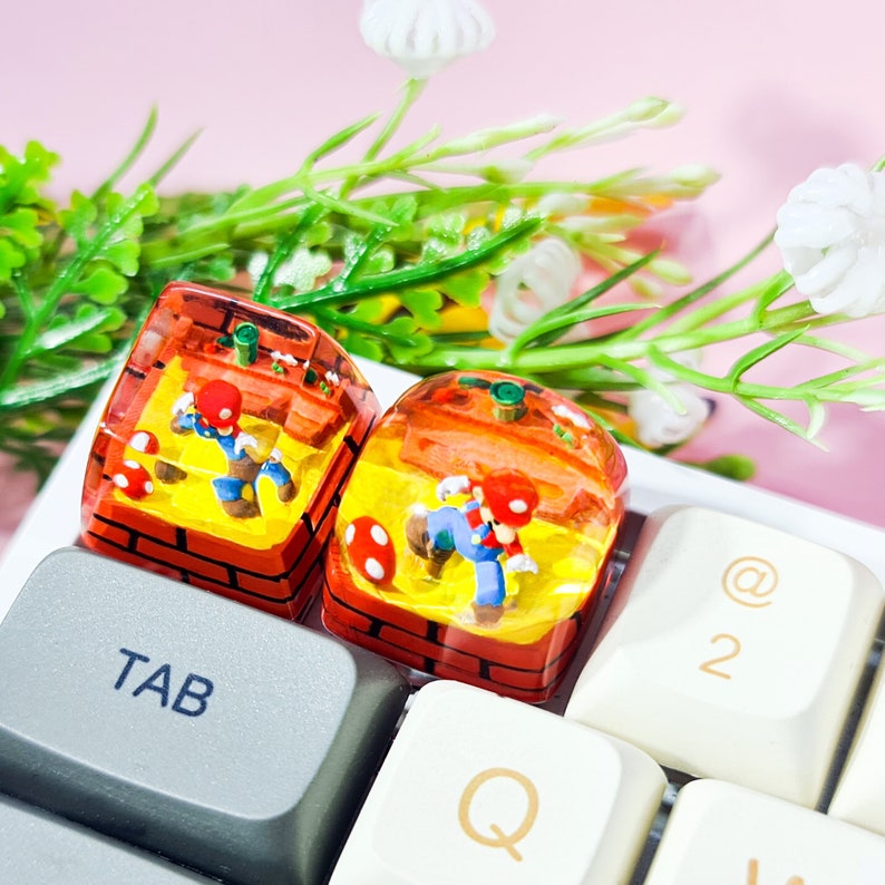 May include: Two resin keycaps featuring a video game character in a brick setting. The keycaps are on a white keyboard with the keys "TAB", "Q", "@", and "2" visible.