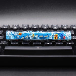 May include: A custom artisan keycap with a blue and white design, depicting cartoon fish and water. The rectangular keycap is set on a black keyboard, adding a unique touch to a mechanical keyboard.
