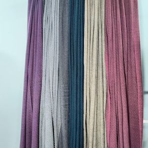 Puede incluir: Exhibición de seis bufandas drapeadas en varios colores, incluyendo morado, gris, verde azulado y beige. Las bufandas tienen un patrón de espiga texturizado. Cuelgan de un soporte blanco contra una pared azul claro.