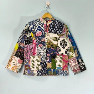 Puede incluir: Chaqueta patchwork con una variedad de estampados florales y geométricos en tonos azules, rosas, verdes y amarillos. La chaqueta tiene cuello mandarín, dos bolsillos delanteros y está colgada de una percha de madera sobre un fondo azul claro.