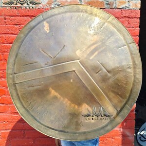 May include: A large, round, antique-style shield with a bronze finish. The shield features a prominent V-shaped design and horizontal lines, with a circular border. The shield is displayed against a red brick wall.