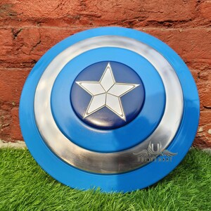 May include: A blue and silver Captain America shield with a white star in the centre. The shield is made of metal and has a raised edge. The shield is on a green grassy surface.