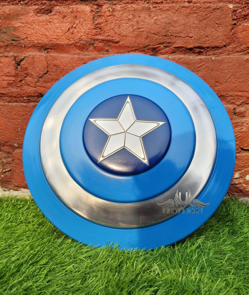 May include: A blue and silver Captain America shield with a white star in the centre. The shield is made of metal and has a raised edge. The shield is on a green grassy surface.