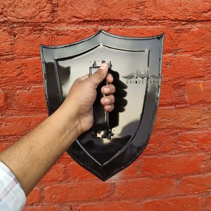Pode incluir: Um escudo preto com acabamento brilhante, segurado contra uma parede de tijolos vermelhos. O escudo tem uma base pontiaguda e uma al&ccedil;a na parte de tr&aacute;s. O texto "UNIQUE ART" &eacute; vis&iacute;vel no escudo.