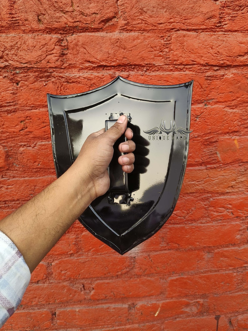 Pode incluir: Um escudo preto com acabamento brilhante, segurado contra uma parede de tijolos vermelhos. O escudo tem uma base pontiaguda e uma al&ccedil;a na parte de tr&aacute;s. O texto "UNIQUE ART" &eacute; vis&iacute;vel no escudo.