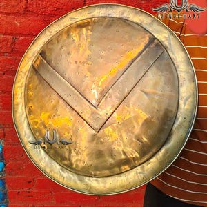 May include: A large, round, antique-style shield with a brass finish. The shield features a prominent V-shaped design in the center and a decorative border. The shield is likely a replica or prop.