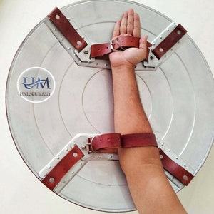 Op de afbeelding: Een zilveren metalen schild met een bruine leren riem en gesp. Het schild heeft een cirkelvormig ontwerp met een verhoogd midden en een stervormig patroon rond de rand. De riem is met metalen klinknagels aan het schild bevestigd en is ontworpen om op de arm te worden gedragen.