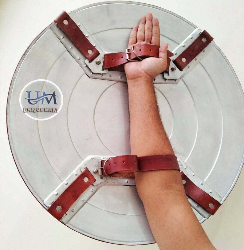 Op de afbeelding: Een zilveren metalen schild met een bruine leren riem en gesp. Het schild heeft een cirkelvormig ontwerp met een verhoogd midden en een stervormig patroon rond de rand. De riem is met metalen klinknagels aan het schild bevestigd en is ontworpen om op de arm te worden gedragen.