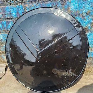 May include: A black, round shield with a V-shaped design. The shield has a glossy finish and a decorative emblem near the bottom. The text "UNIQUE U MAAT" is visible at the top. This item is likely a decorative piece or a costume accessory.