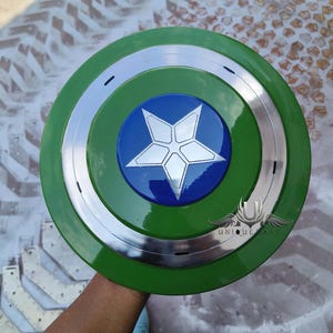May include: A green and silver Captain America shield with a blue star in the centre. The shield has a raised rim and a smooth surface. The star has a white outline and a five-pointed design. The shield is held by a hand in the centre of the image. The text "UNIQUE MART" is visible in the lower right corner of the image.