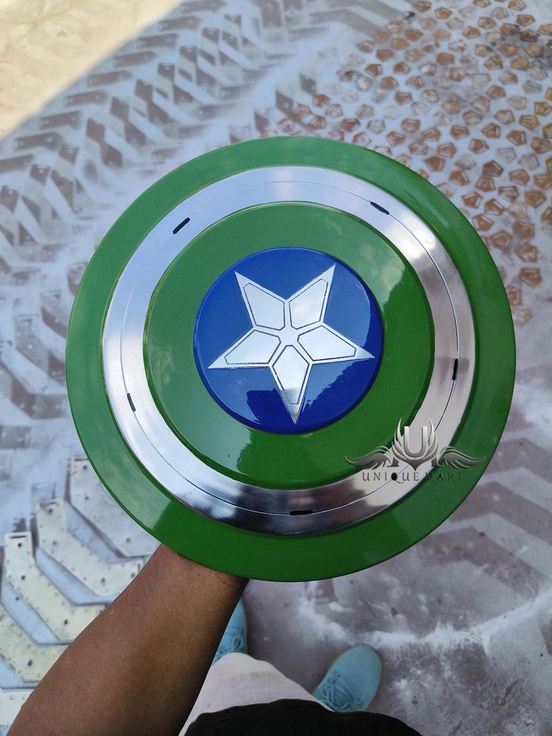 May include: A green and silver Captain America shield with a blue star in the centre. The shield has a raised rim and a smooth surface. The star has a white outline and a five-pointed design. The shield is held by a hand in the centre of the image. The text "UNIQUE MART" is visible in the lower right corner of the image.