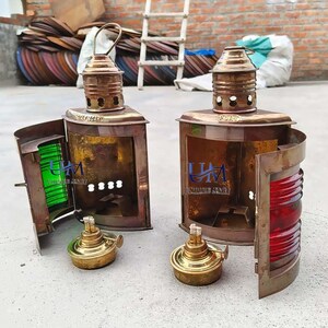 Pu&ograve; includere: Due antiche lanterne nautiche in ottone con pannelli di vetro e inserti in vetro rosso e verde. Le lanterne hanno una porta a battente e una maniglia in ottone sulla parte superiore. Le lanterne sono su una superficie di cemento.
