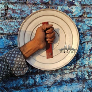 May include: A silver round shield with a red leather handle. The shield has a raised rim and is made of metal. The handle is attached to the back of the shield and has a small metal loop at the top.