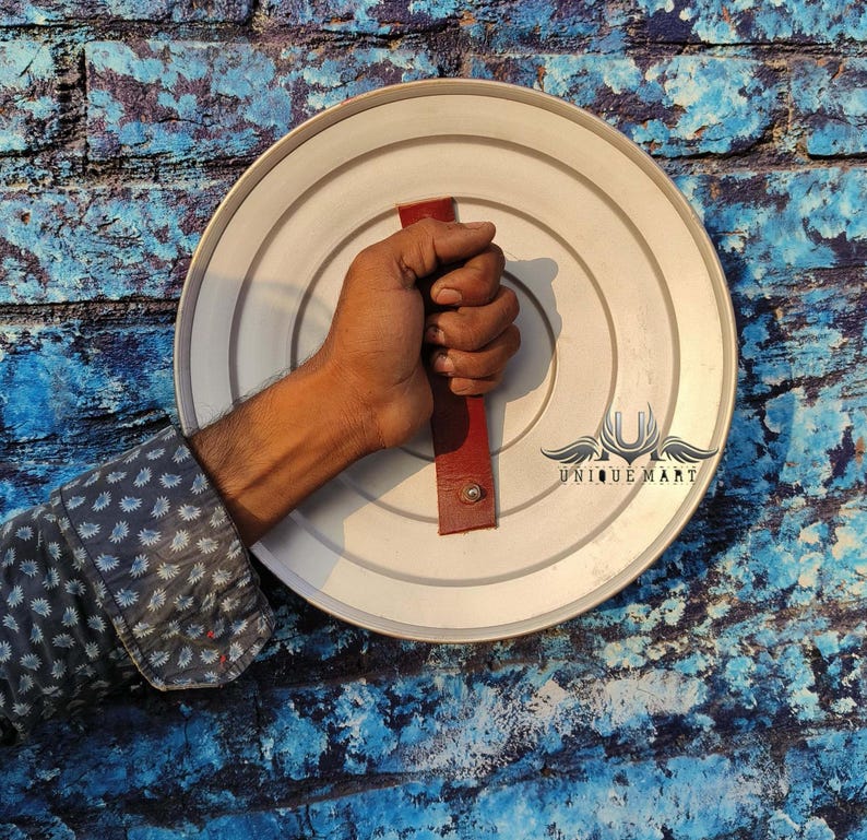 May include: A silver round shield with a red leather handle. The shield has a raised rim and is made of metal. The handle is attached to the back of the shield and has a small metal loop at the top.