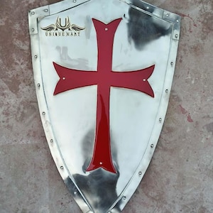 May include: A silver shield with a red cross in the centre. The shield has a rounded top and a pointed bottom. The shield is decorated with rivets around the edge.