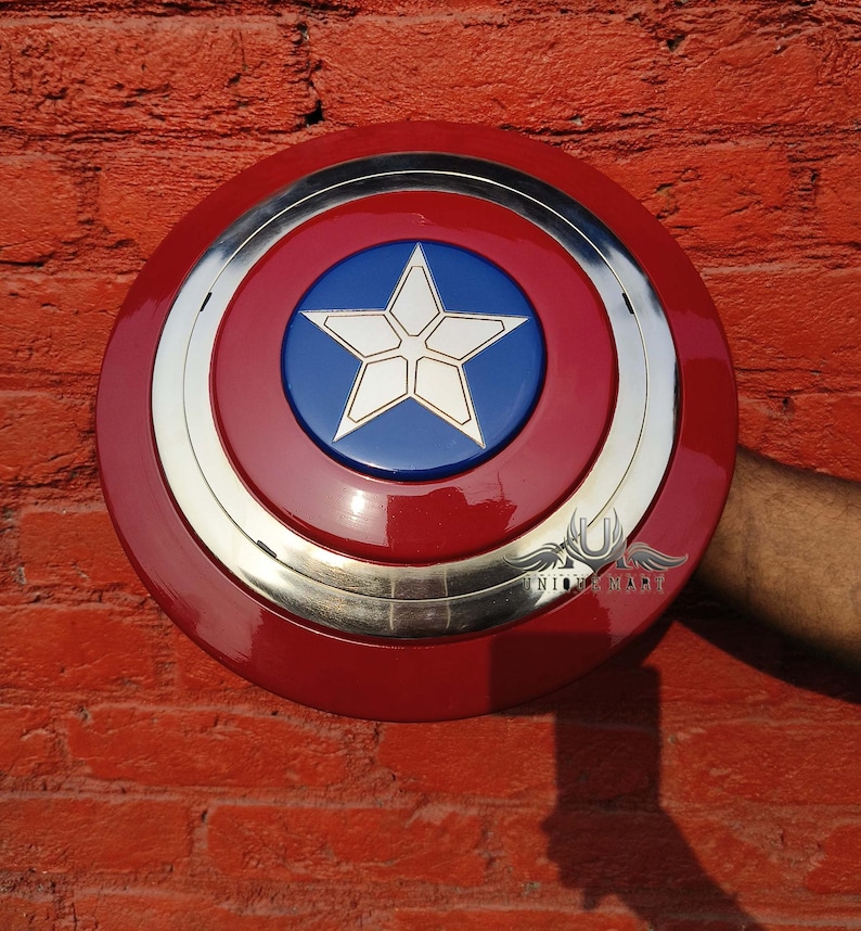 May include: A red, white and blue Captain America shield with a silver metal rim. The shield is held in a hand against a red brick wall.
