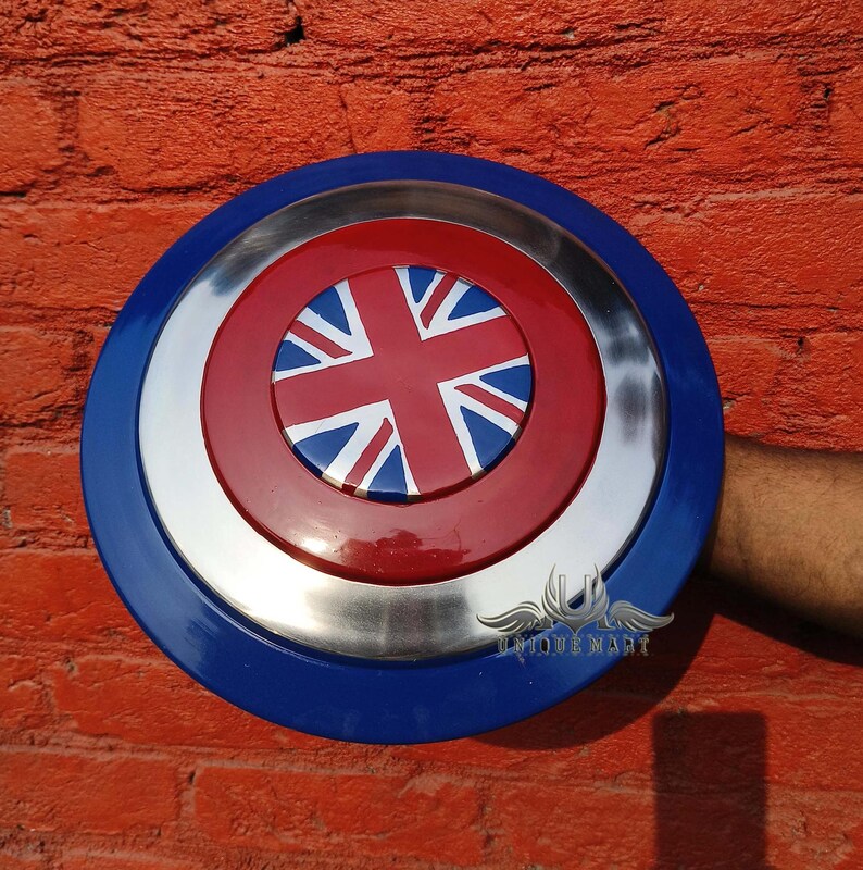 May include: A blue, red and white shield with a Union Jack design in the centre. The shield has a silver metal rim and is being held by a hand.