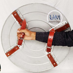 Op de afbeelding: Een zilveren metalen schild met een rode leren riem en gesp. Het schild heeft een ontwerp met een driepuntige ster en wordt vastgehouden door een hand die een zwart shirt draagt. Het schild heeft de tekst "UM UNIQUE MART" erop.