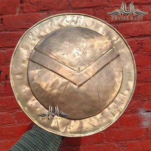 May include: A round, antique-style shield with a bronze finish. The shield features a prominent chevron design and a circular outer rim. The words "UNIQUE MART" are visible in the upper right and lower center of the shield.
