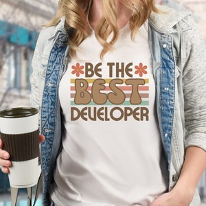 May include: A white t-shirt with a retro-style graphic that says "*BE THE* BEST DEVELOPER" with a flower on either side of the text. The graphic is in brown, orange, and yellow with a striped background.