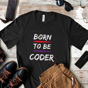 May include: Black t-shirt with the text "BORN TO BE CODER" in colorful lettering. The shirt is laid out on a wooden surface with a brown leather bag, sunglasses, and brown pants.