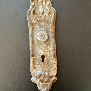 May include: An antique, ornate door plate with a keyhole and a decorative, weathered finish. The metal plate has a rectangular shape with detailed floral and scrollwork designs at the top and bottom. The center features a hexagonal detail around the knob.