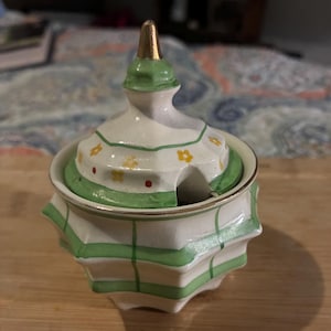 May include: A white ceramic sugar bowl with a faceted, star-shaped body featuring green horizontal and vertical stripes. The matching lid has a gold-toned finial, green trim, and a pattern of small yellow flowers and red dots with a spoon notch.
