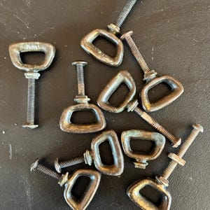 May include: A collection of antique bronze-colored D-ring pulls with threaded bolts. The pulls are in various orientations, showing signs of age and wear. The hardware is likely for furniture or cabinetry, offering a vintage aesthetic.