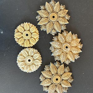 May include: Five antique brass-colored floral decorative elements. Each piece features a central hole surrounded by detailed leaves and small rose accents. The pieces vary slightly in color and finish, with a vintage aesthetic.
